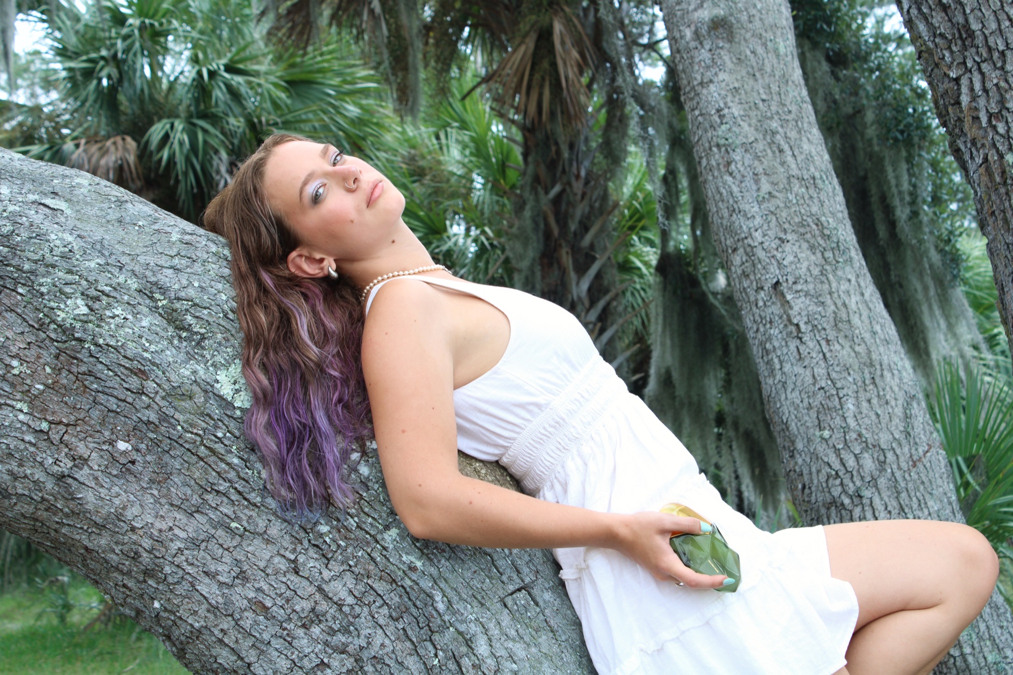Film still — the model reclining in a live oak draped with Spanish moss, holding the Salted Muse bottle. The Garden of You as a real place.